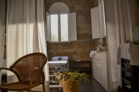 Sala de tratamiento con muros de piedra, camilla y butaca de mimbre en el Gran Balneario do Carballiño